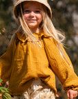 Child in a yellow outfit with a hat, standing outdoors among plants.