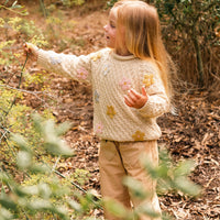 Child in a forest wearing a light-colored sweater and pants, surrounded by greenery.