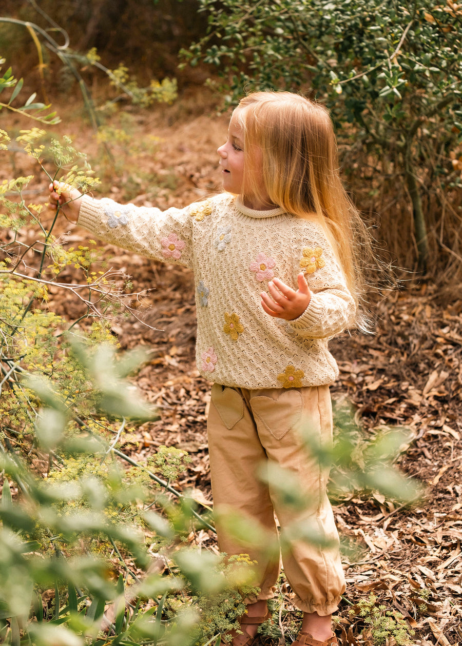 Child in a forest wearing a light-colored sweater and pants, surrounded by greenery.