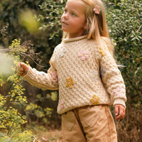 Young girl in a beige sweater and brown pants standing in a forest setting